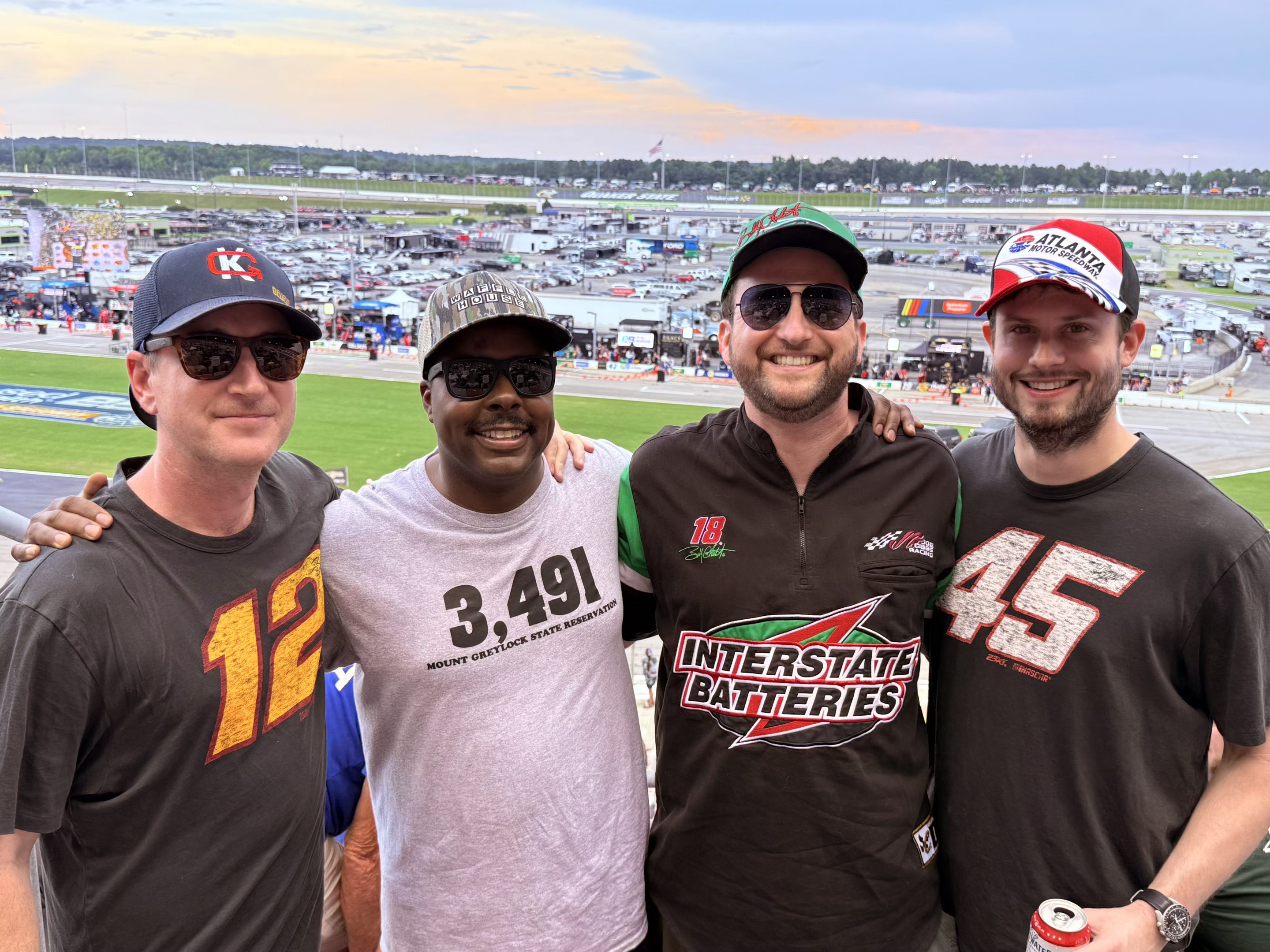 Four thirtysomething men in caps and short-sleeve shirts, all but one of whom are wearing sunglasses, standing arm in arm in front of a race track